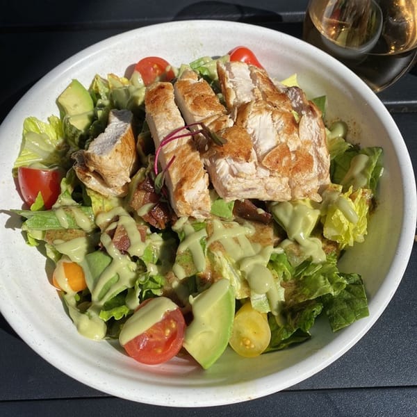 A healthy Good Kitchen salad with grilled chicken, avocado, tomatoes, and green dressing in a white bowl.