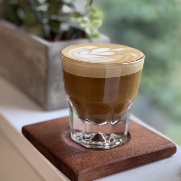 A latte with intricate foam art sits on a wooden coaster by a window at Good Kitchen.