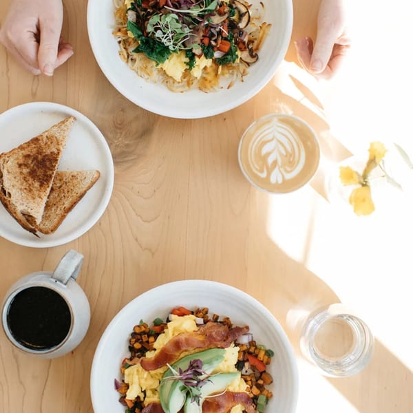 Two healthy breakfast bowls, coffee, and toast on a sunlit wooden table at Good Kitchen.